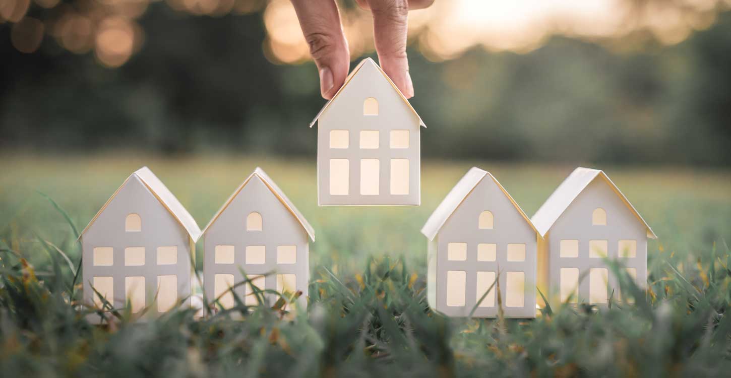 hand choosing white paper house model from group of houses on green grass